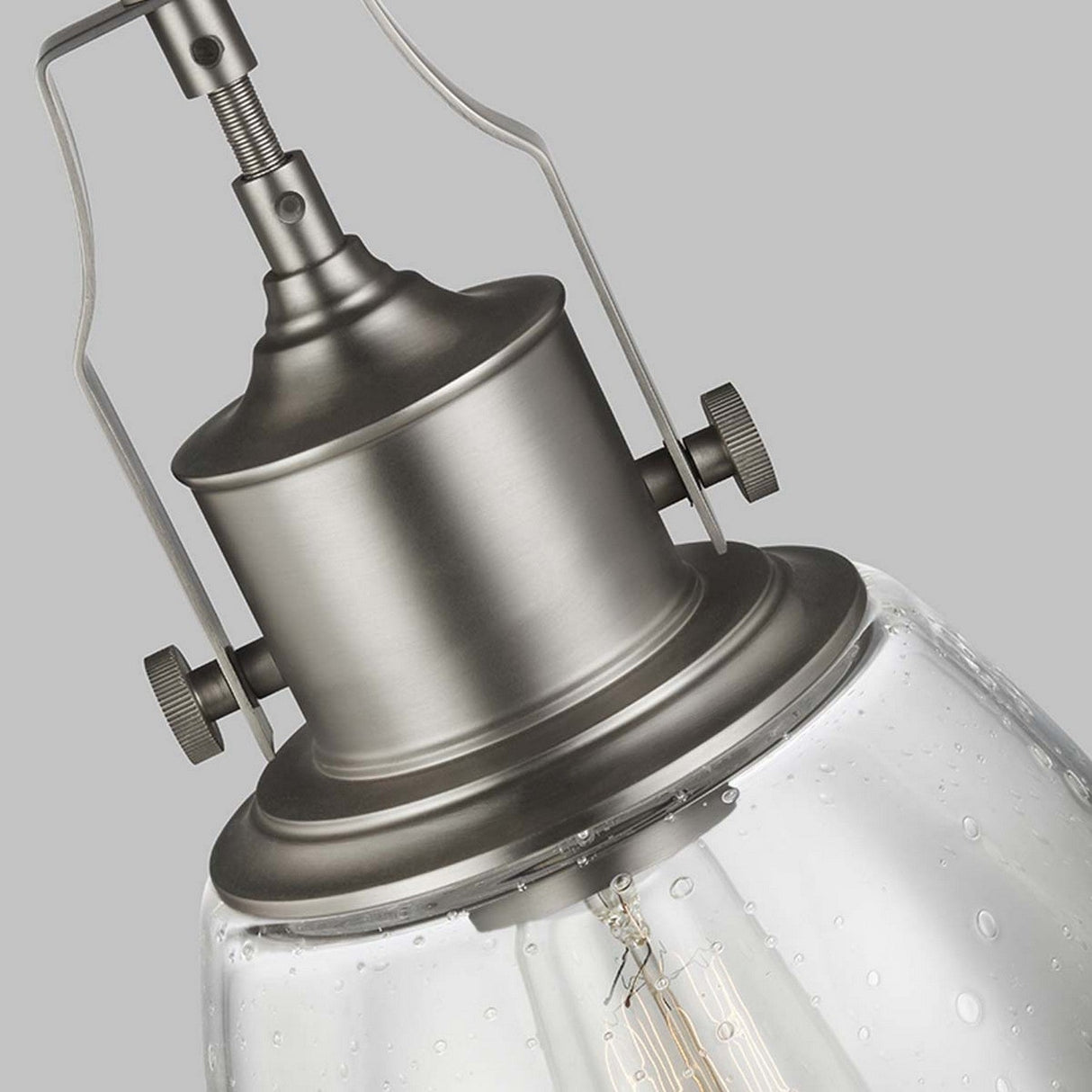 A close-up view of the Hobson 1 Light Medium Pendant in satin nickel reveals its vintage-style design with a clear glass shade that showcases an exposed filament bulb inside. The pendant light, against a plain gray background, highlights its industrial-inspired aesthetic.