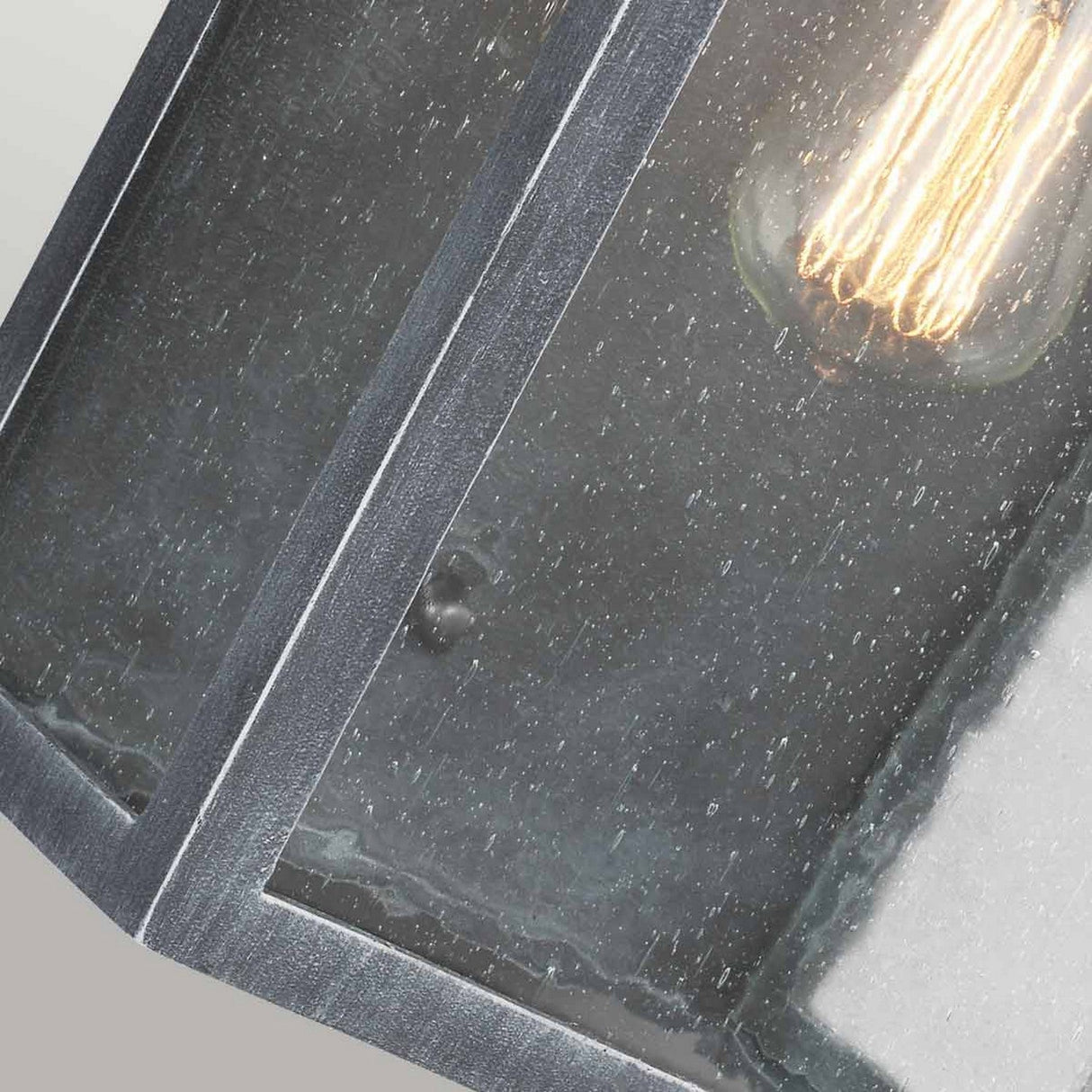 Close-up of the Hodges Outdoor Large Wall Lantern in Ash Black, showcasing its vintage-style charm. The clear seeded glass panels resemble rain-speckled surfaces, while the bulb casts a warm glow, crafting a rustic and cozy atmosphere reminiscent of prairie design.