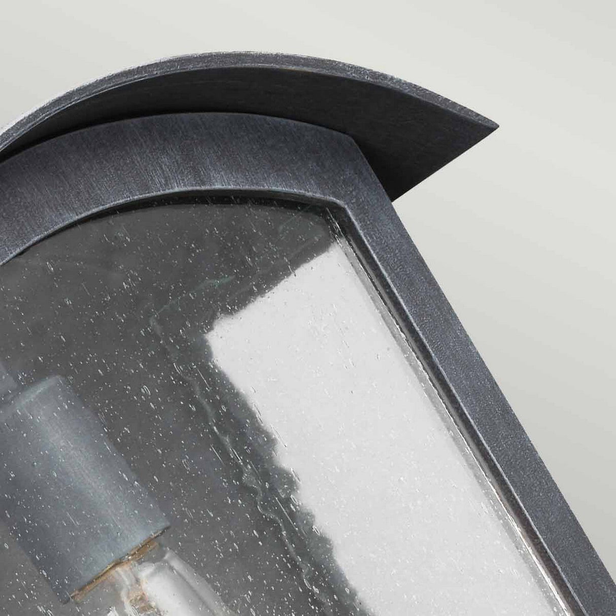 Close-up image of the Hodges Outdoor Large Wall Lantern in ash black, featuring a curved top edge and clear glass panel with raindrop details that partially reveal the light bulb inside, enhancing the farmhouse aesthetic.
