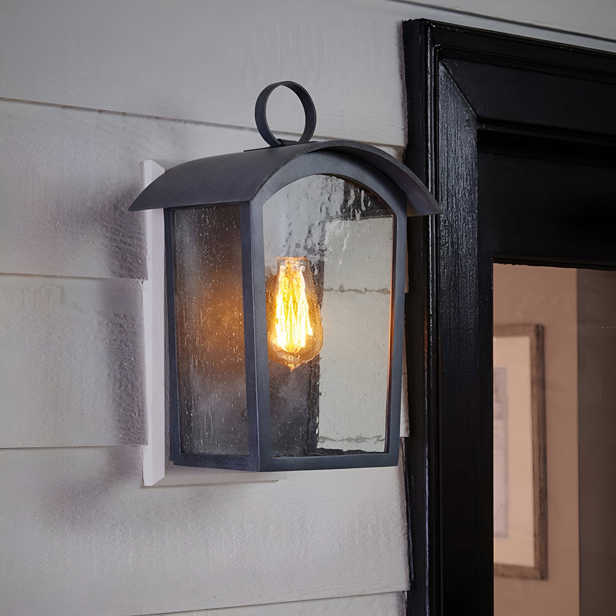 A Huguenot Lake 1 Light Wall Light in polished chrome is mounted on a white wall beside a doorframe. The warm, glowing bulb within its black metal and glass enclosure casts an inviting glow.