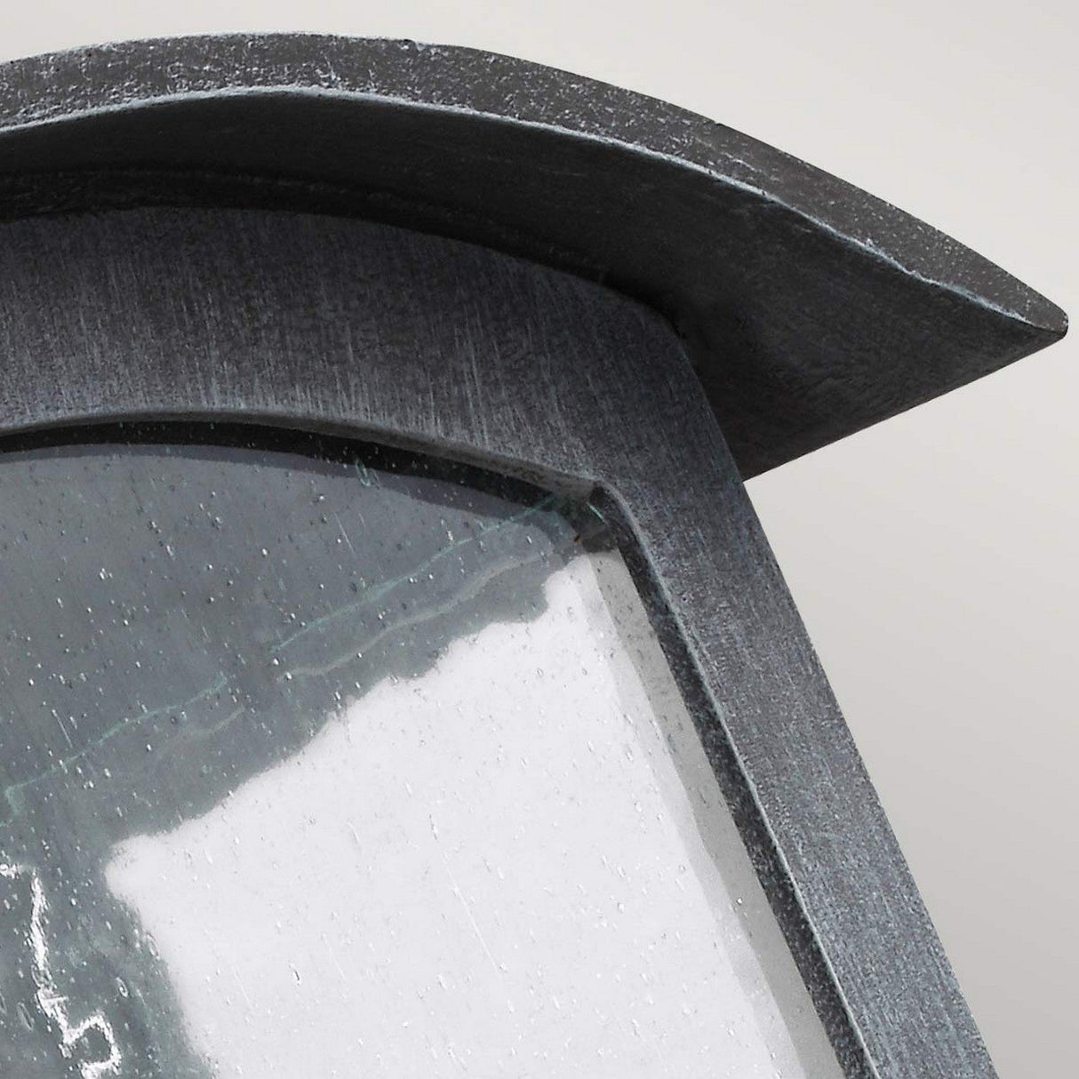 Close-up of a rain-drenched Hodges Outdoor Small Wall Lantern - Ash Black, showcasing its curved glass window and dark metallic frame. Raindrops streak the glass, evoking a prairie design aesthetic against a soft, blurred gray background.