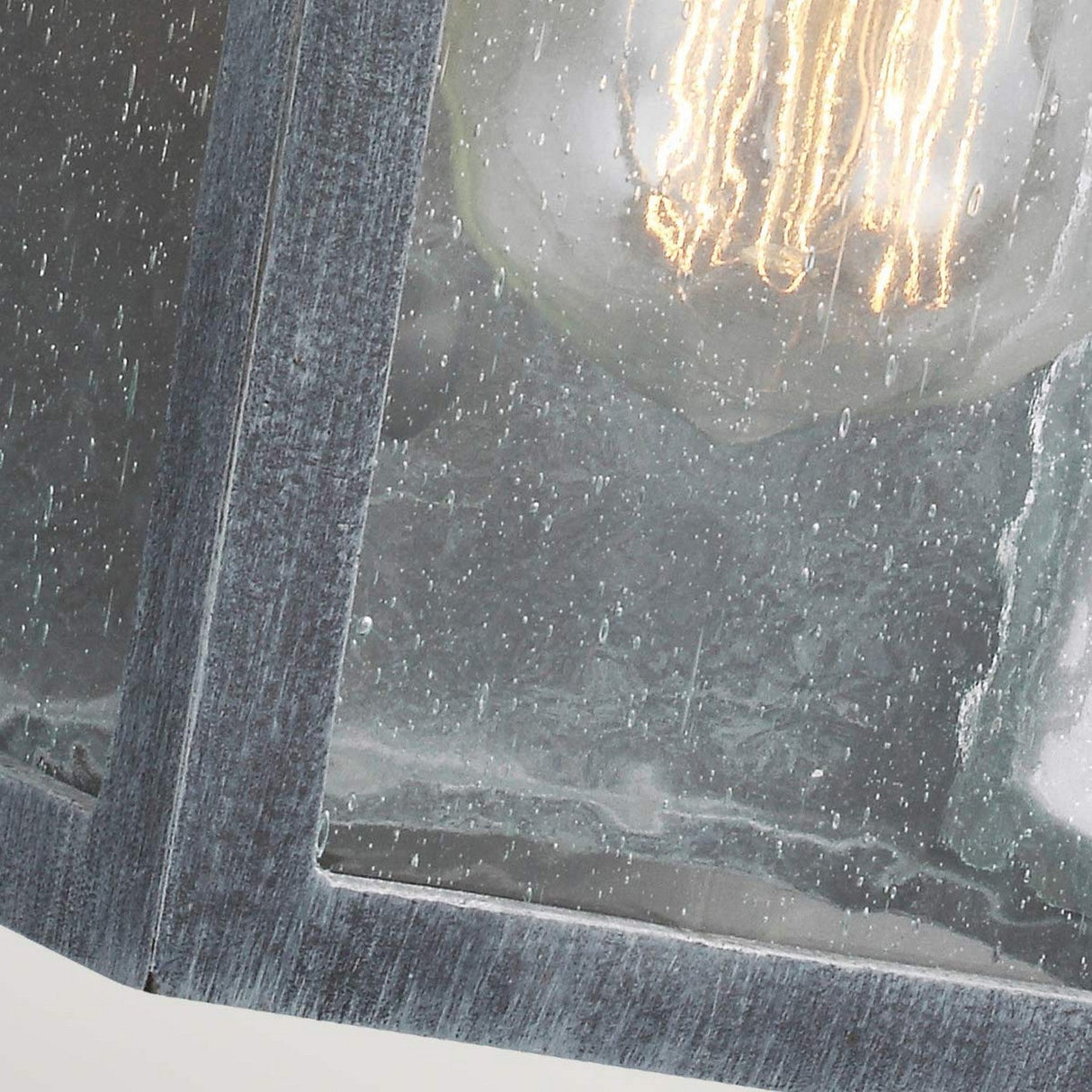 Close-up of the Hodges Outdoor Small Wall Lantern in Ash Black, with raindrops on the glass. Behind the pane, a glowing filament bulb casts a warm light, creating a prairie design that contrasts with the gray and wet surface of its frame.
