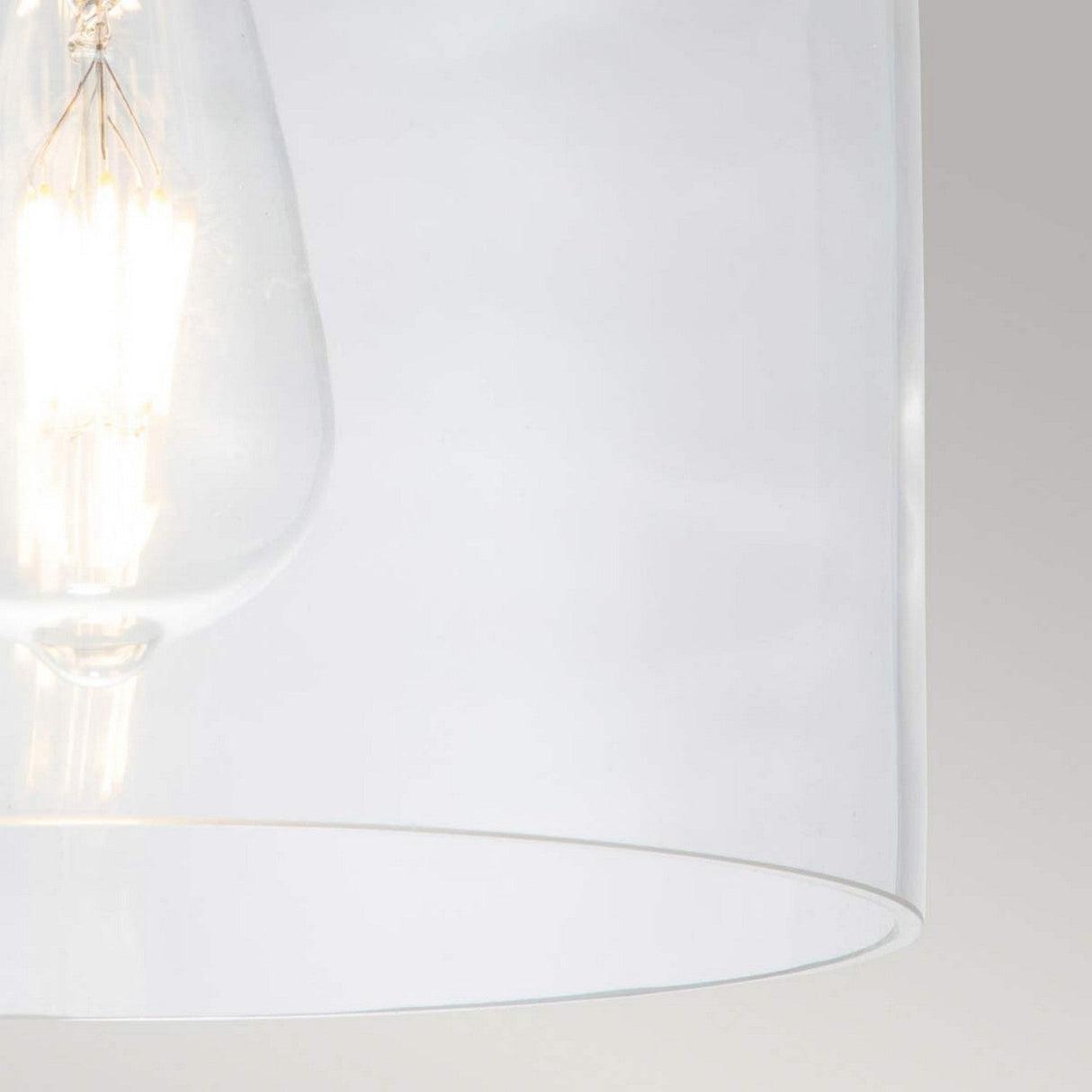 Close-up of a clear glass light bulb glowing warmly inside the Hounslow 1 Light Flush - Burnished Brass against a plain light gray background. The image highlights the bulb's filament and modern design, complemented by subtle burnished brass accents for an elegant touch.