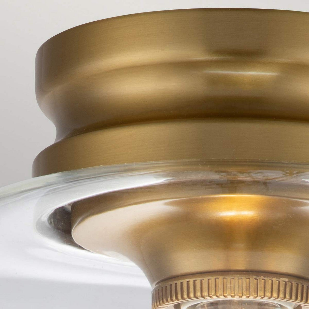 Close-up of the Hounslow 1 Light Flush - Burnished Brass ceiling light fixture, featuring a circular design. The metal showcases a polished finish, complemented by a clear glass rim around the base for a modern and elegant appearance.