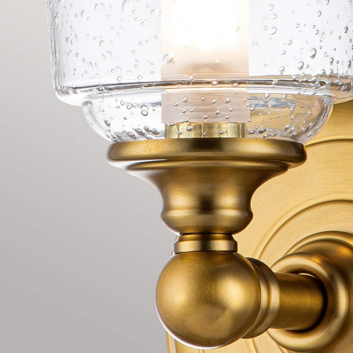 A detailed view of the Huguenot Lake 1 Light Wall Light in Burnished Brass, showcasing a seeded glass shade. When illuminated, it emits a cozy glow, highlighting its elegant and traditional design with graceful curves and a polished surface.
