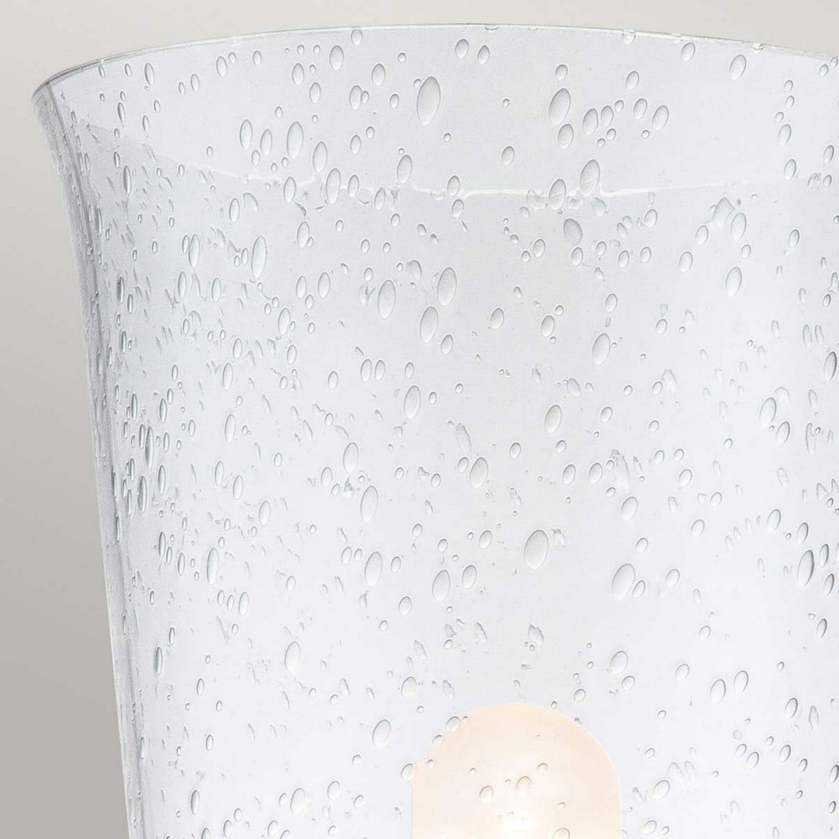A close-up of the Huguenot Lake 1 Light Wall Light reveals its frosted, seeded glass lampshade featuring a raindrop pattern. The light is turned on, casting a soft glow through the textured surface and highlighted by the elegant burnished brass accents.