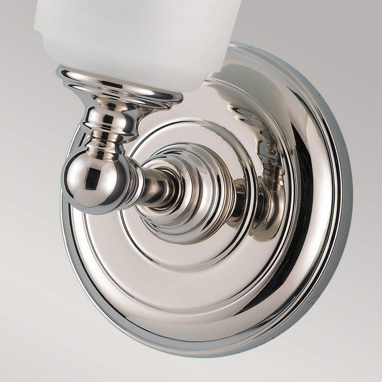 Close-up of the Huguenot Lake 1 Light Wall Light in polished chrome, showcasing its ornate, circular base. The wall light features a curved arm that supports an opal glass shade, offering modern and elegant bathroom lighting against a plain background.