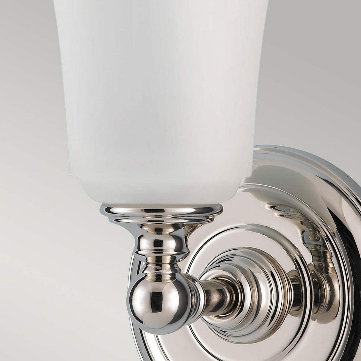 The Huguenot Lake 1 Light Wall Light in polished chrome, with its opal glass shade, enhances the bathroom lighting. Its decorative round base and sleek, modern design stand out against a soft gray background.