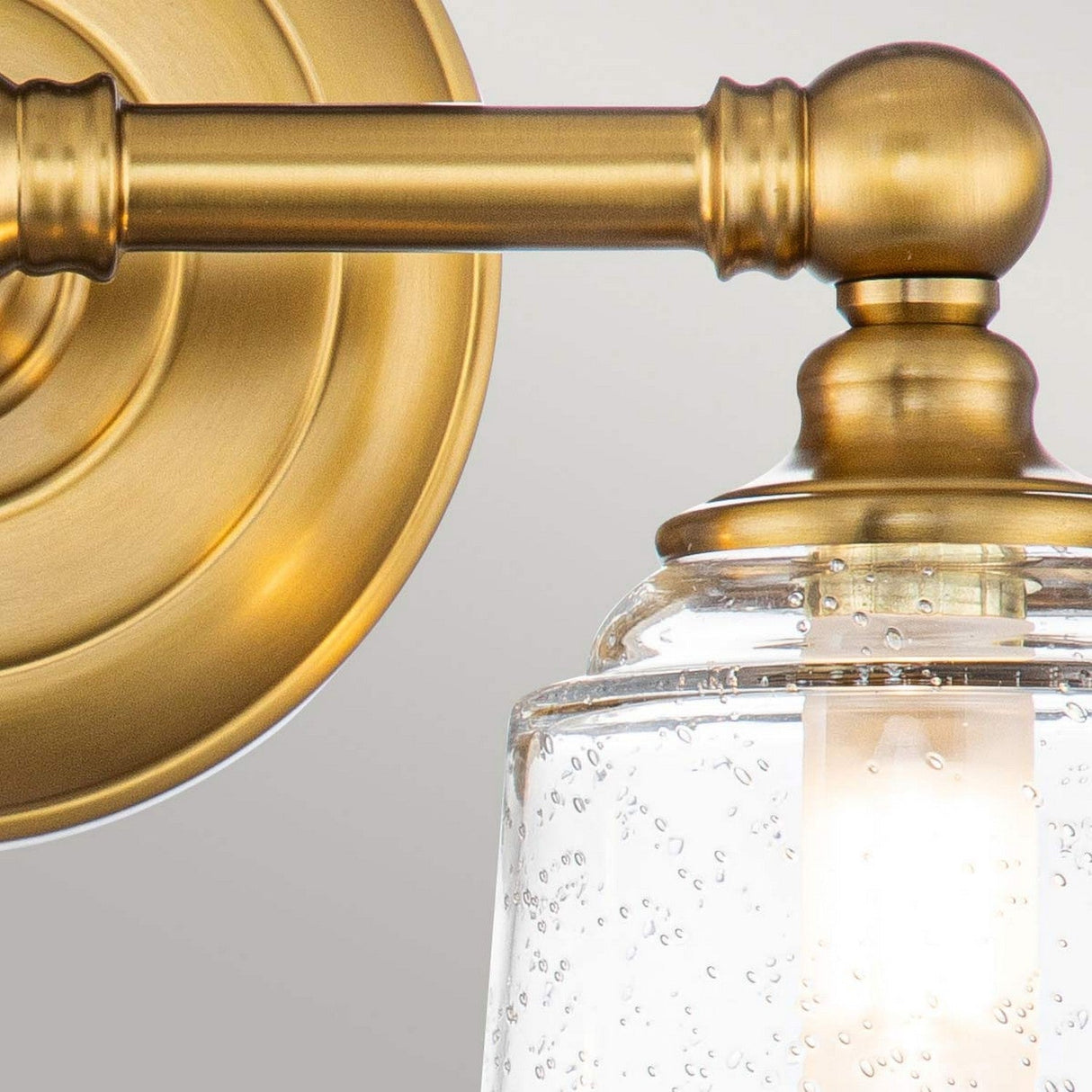 Close-up of the Huguenot Lake 2 Light Wall Light in burnished brass, featuring a clear, seeded glass shade. The light is illuminated, casting a warm glow, and is affixed to a circular brass base.