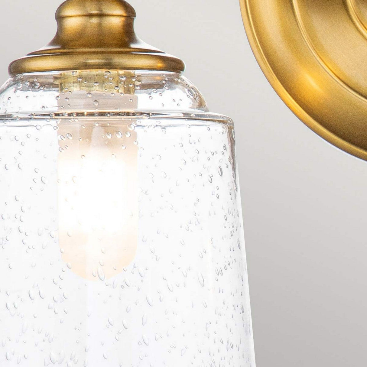 A close-up of the Huguenot Lake 2 Light Wall Light in a burnished brass finish reveals its clear, seeded glass shade that encases a lit bulb, emitting a soft glow and adding an elegant touch to the design.