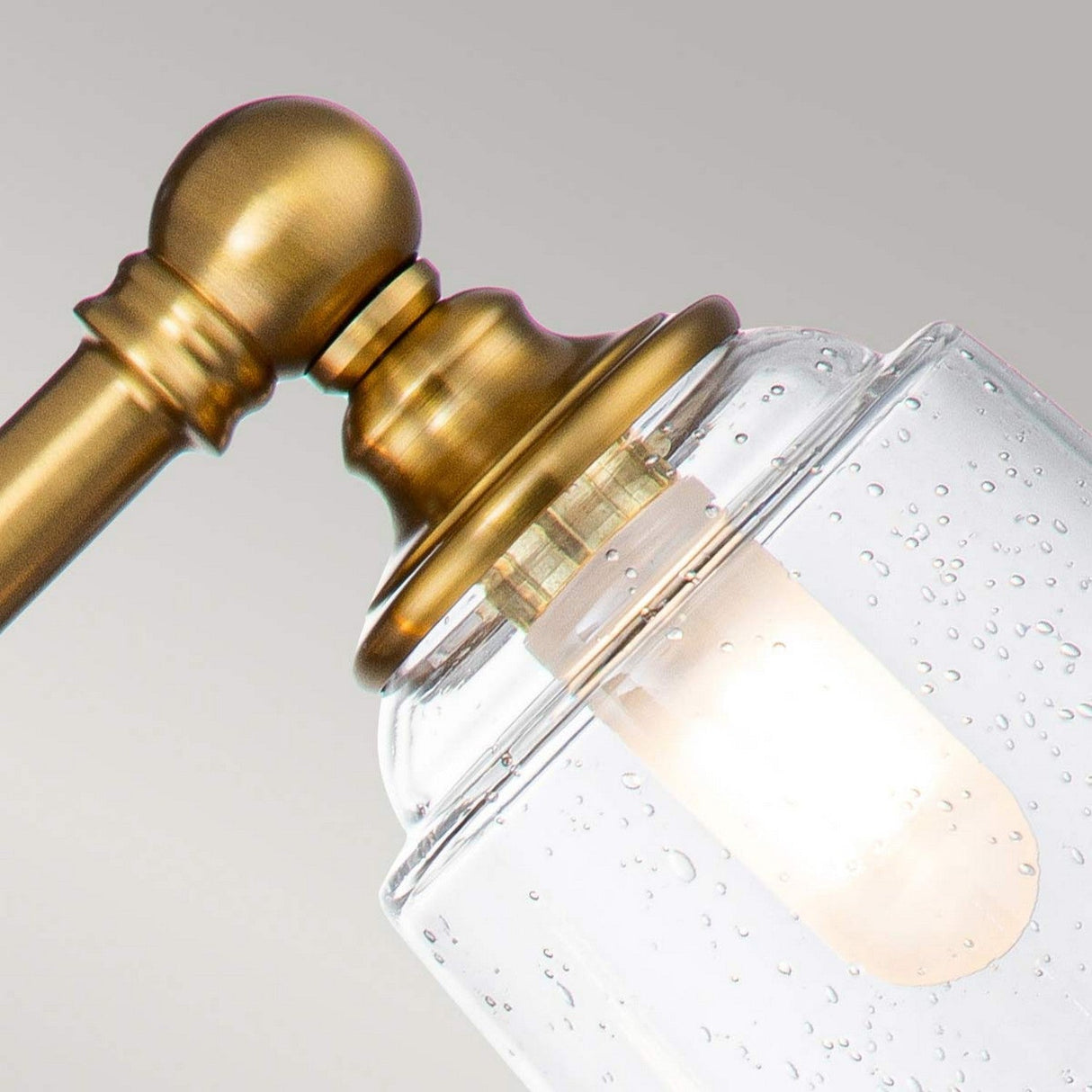 A close-up of the Huguenot Lake 3 Light Wall Light in burnished brass, featuring a clear, seeded glass shade with a lit bulb inside. The glass is speckled with small air bubbles, enhancing the elegant Colonial design against a plain, neutral background.