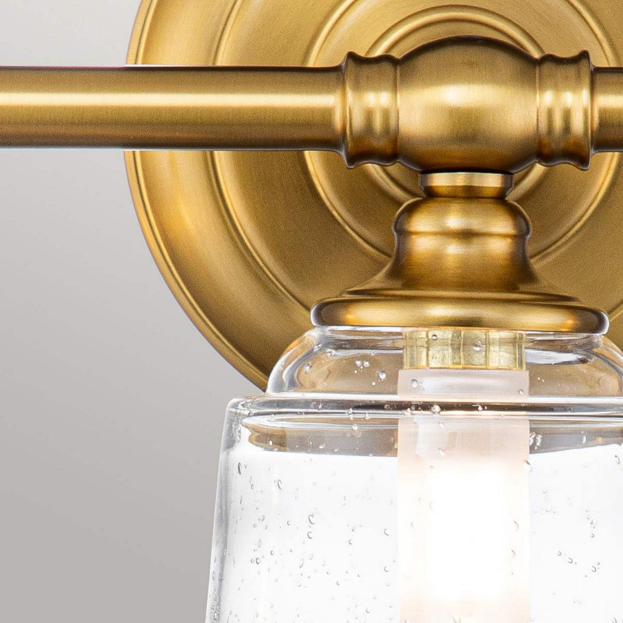 Close-up of the Huguenot Lake 3 Light Wall Light in burnished brass, highlighting its elegant curves and clear, seeded glass shade. The colonial design features a warm metallic finish with light gently glowing through the textured glass.