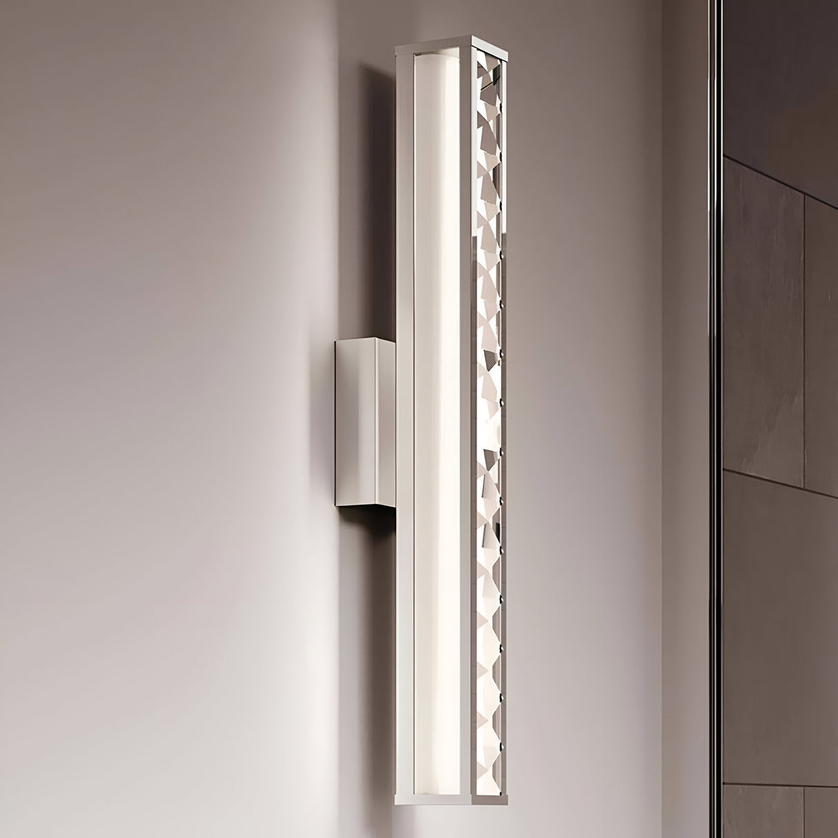 The Jessie 1 Light LED Wall Light, with its rectangular design and polished chrome finish, features a patterned side panel. The light softly illuminates a neutral-colored wall, crafting an elegant and contemporary ambiance.