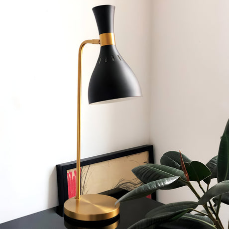 The Joan 1 Light Table Lamp in Midnight Black and Burnished Brass sits sleekly on a black surface beside a large green plant. A framed artwork with an abstract design rests flat against the white wall behind this modern lamp.