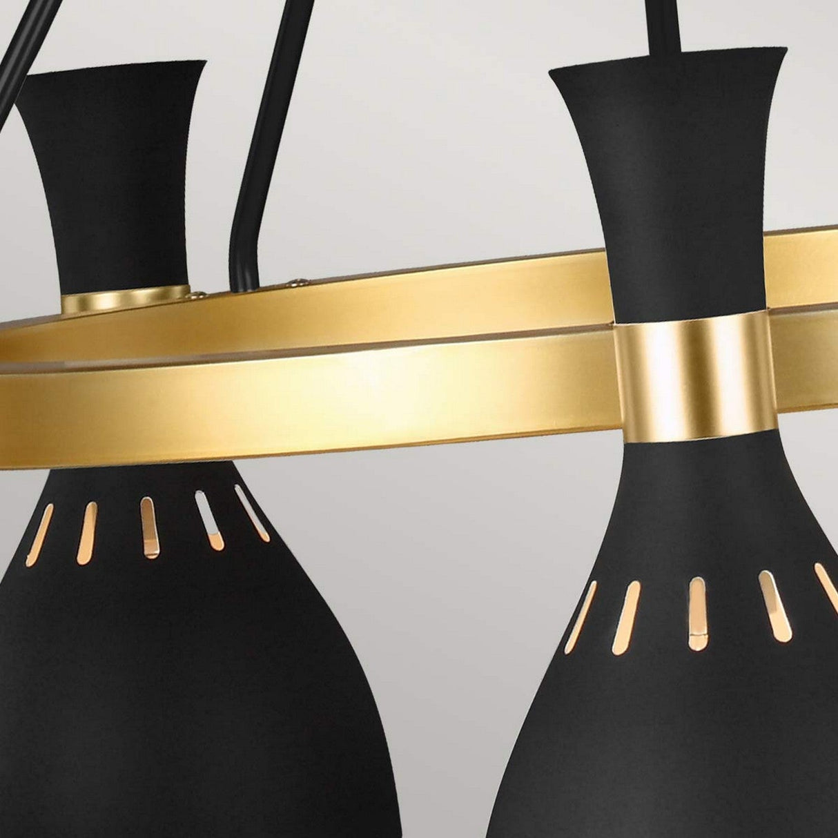 A close-up of the Joan 5 Light Chandelier - Midnight Black/Burnished Brass highlights its conical pendant lights with intricate cutout details. These lights elegantly hang from a circular frame in burnished brass, set against a plain soft gray backdrop.