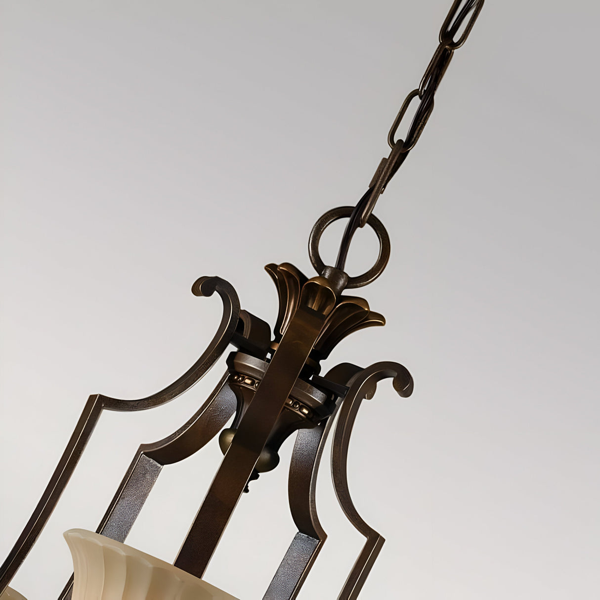 A close-up of the Kelham Hall 5 Light Up Chandelier in Firenze Gold/British Bronze showcases its intricate metalwork, suspended by a decorative chain. The fixture features detailed embellishments and cream-colored glass shades, set against a neutral background.