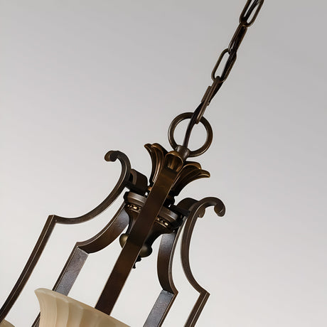 A close-up of the Kelham Hall 5 Light Up Chandelier in Firenze Gold/British Bronze showcases its intricate metalwork, suspended by a decorative chain. The fixture features detailed embellishments and cream-colored glass shades, set against a neutral background.