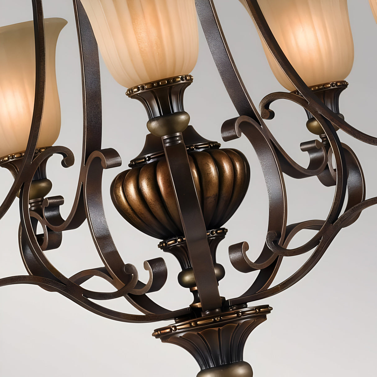 A close-up of the Kelham Hall 5 Light Up Light Chandelier highlights its exquisite design, comprising three elegant antique brass arms and frosted glass shades. With ornate scrollwork and a ribbed central column, this chandelier exudes classic sophistication in its distinctive British Bronze finish complemented by Firenze Gold.