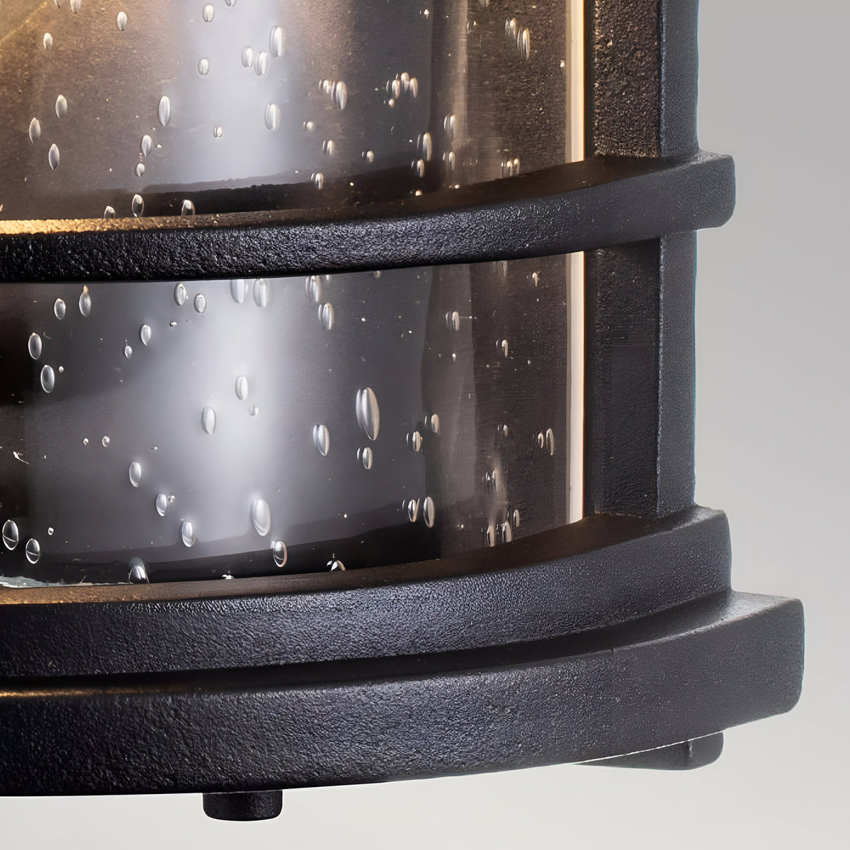 A close-up of the Lighthouse 1 Light Outdoor Half Lantern in Textured Black, featuring a circular and sturdy design with parallel horizontal lines. The lantern's glass enclosure shows condensation droplets while it is lit, casting a warm glow against them.