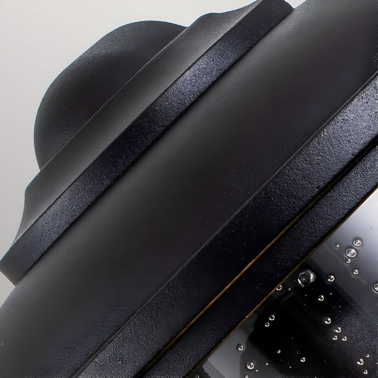 Close-up of a dome-shaped object reminiscent of the Lighthouse 1 Light Outdoor Half Lantern in Textured Black, featuring a smooth texture and glossy finish. Tiny bubbles and droplets of water embellish the lower section, adding a subtly reflective effect. The background is a neutral light gray.