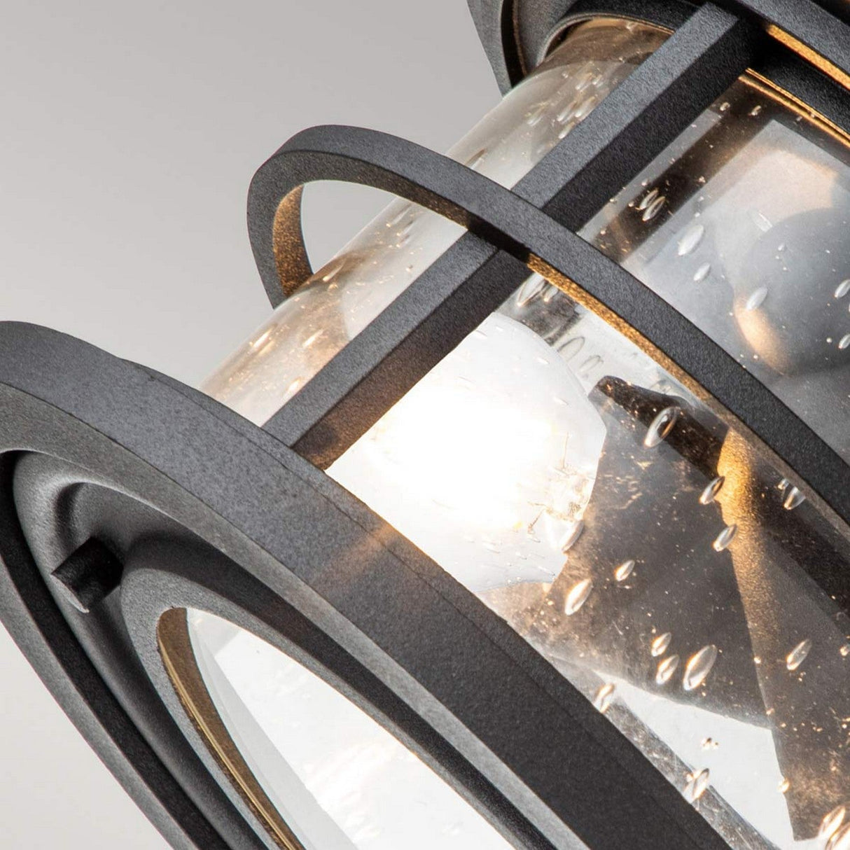 A close-up of the Lighthouse 2 Light Outdoor Flush reveals its modern design, with a clear glass casing surrounded by a textured black metal framework. The light bulb inside provides a warm glow, and small bubbles in the glass enhance the depth and intrigue of the design.