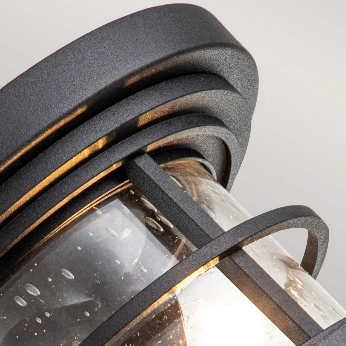 A close-up of the Lighthouse 2 Light Outdoor Flush in textured black reveals its clear glass enclosure. Designed similarly to a black metal lantern, it features layered metal bands and droplets reminiscent of recent rain on the glass surface. Soft light emanates from within, casting shadows across its textured finish.