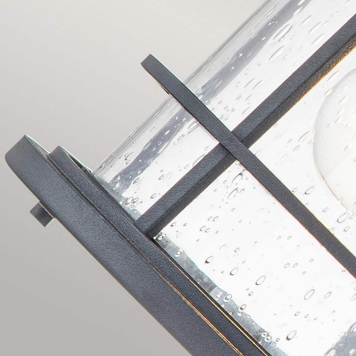 Close-up of the Lighthouse 1 Light Outdoor Medium Wall Lantern in Textured Black, featuring a glass panel adorned with water droplets and enclosed within a nautically inspired black metal frame. The partially lit glass provides both transparency and reflection, set against a soft, neutral gray background.