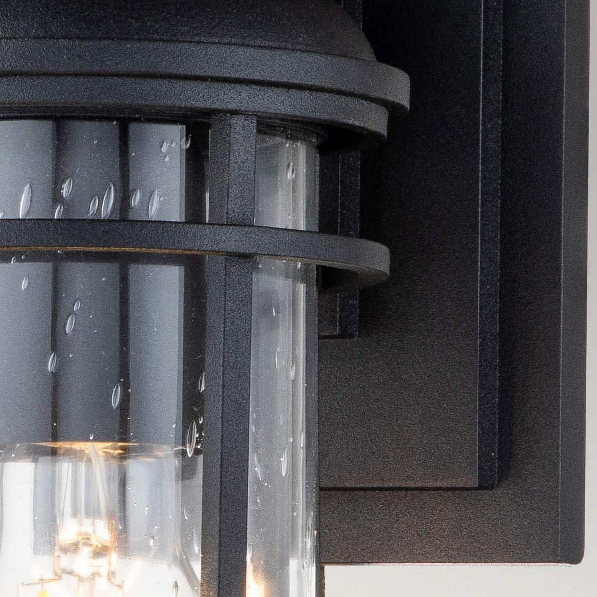 A detailed view of the Lighthouse 1 Light Outdoor Small Wall Lantern in Textured Black showcases its nautical-inspired design, featuring a glass encasement dotted with water droplet textures and a softly glowing bulb that emits a gentle illumination.