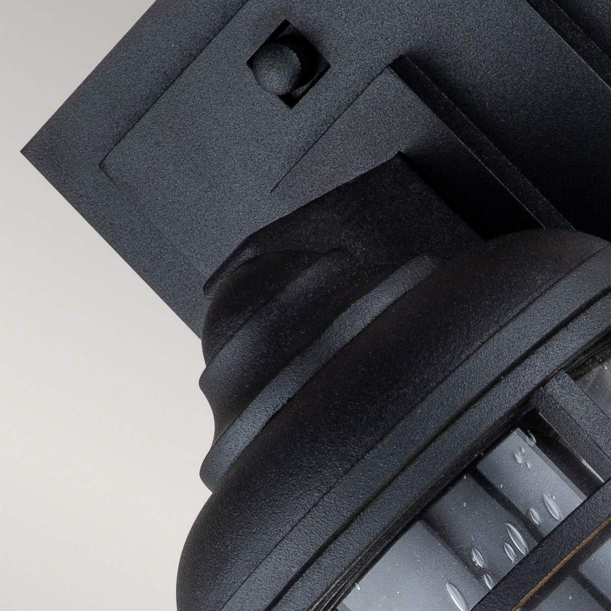 Close-up of a Lighthouse 1 Light Outdoor Small Wall Lantern in textured black, mounted on an outdoor wall against a plain background. The fixture showcases a modern design with a square backing and a rounded light cover adorned with small vertical slits, subtly reflecting its nautical-inspired charm.