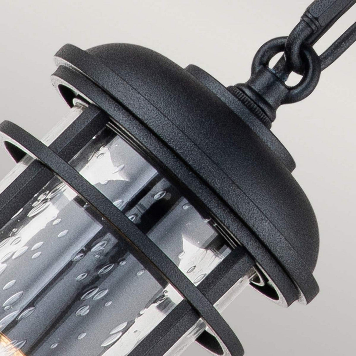 Close-up of the Lighthouse 1 Light Outdoor Small Chain Lantern in a textured black finish with clear seeded glass, highlighting water droplets reminiscent of recent rain or high humidity. The lantern elegantly hangs from a curved hook.