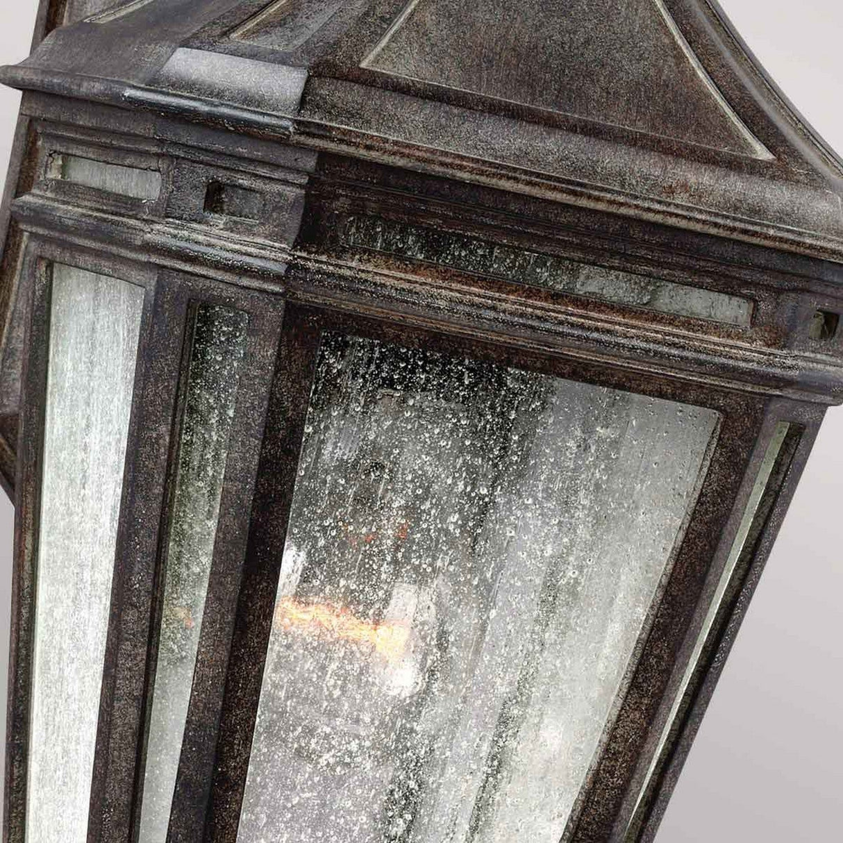 Close-up of the Londontowne 1 Light Outdoor Wall Lantern in weathered chestnut: a vintage-style piece featuring a textured seeded glass enclosure. The metal frame shows signs of rust and patina, while a warm light softly glows inside, casting gentle illumination.
