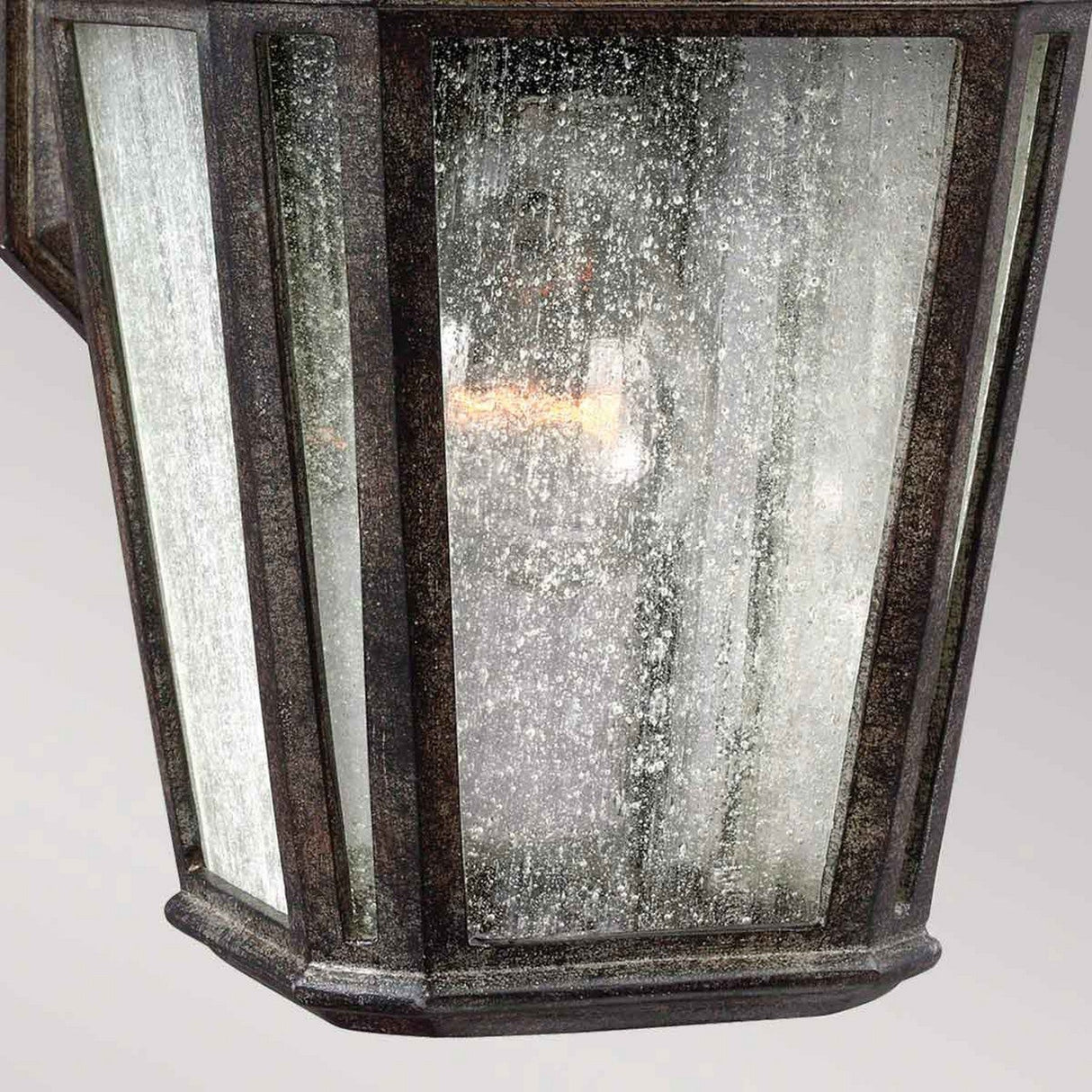 The Londontowne 1 Light Outdoor Wall Lantern in weathered chestnut features a rustic design with speckled, seeded glass panels. Its black frame enhances the antique appearance, and it casts a warm glow from the light bulb within.