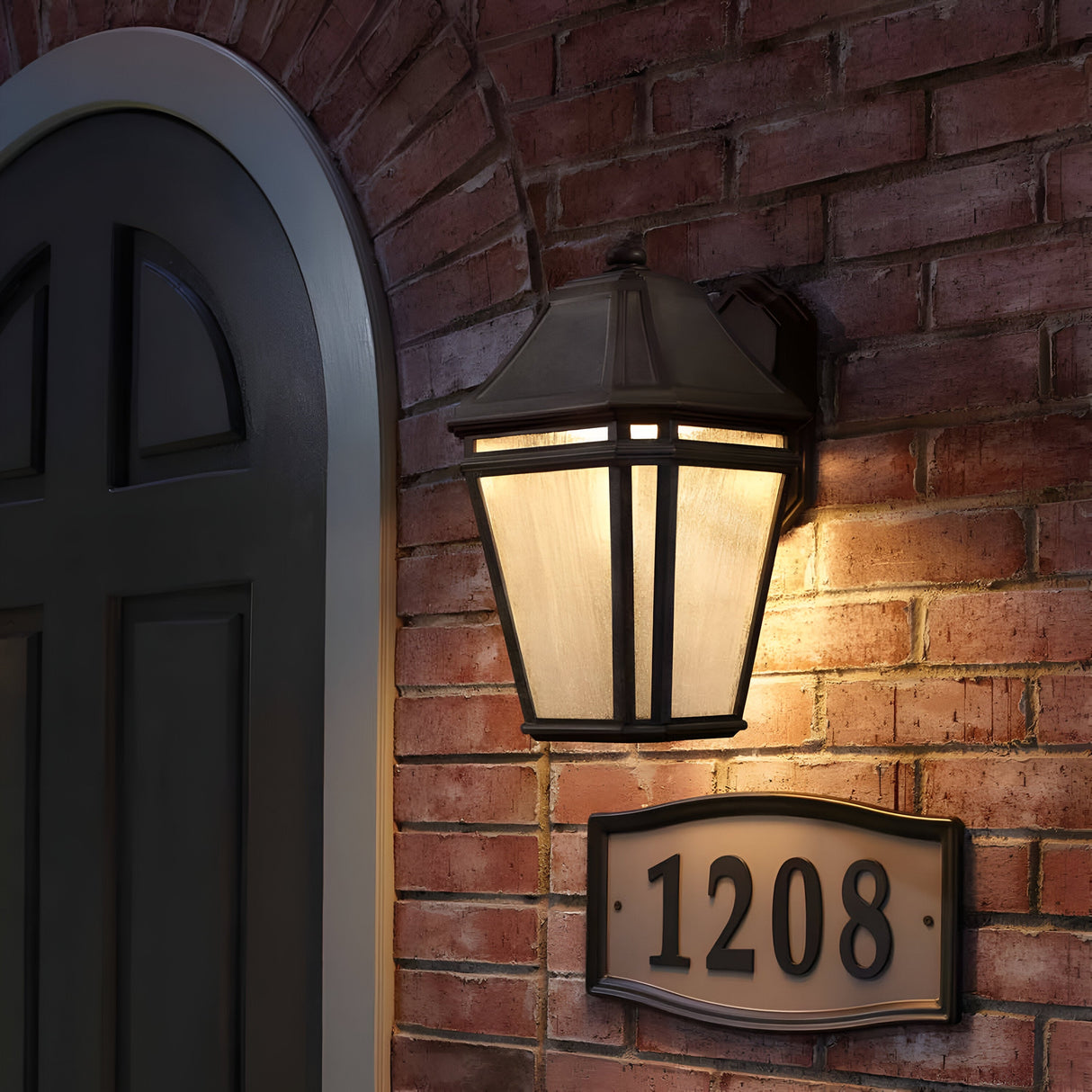 A brick wall featuring an arched doorway on the left displays the Londontowne 1 Light Outdoor Wall Lantern, which emits a warm glow through its seeded glass. Above it, a house number plaque reads 1208. The weathered chestnut finish of the lantern hints at the entrance to a charming home.