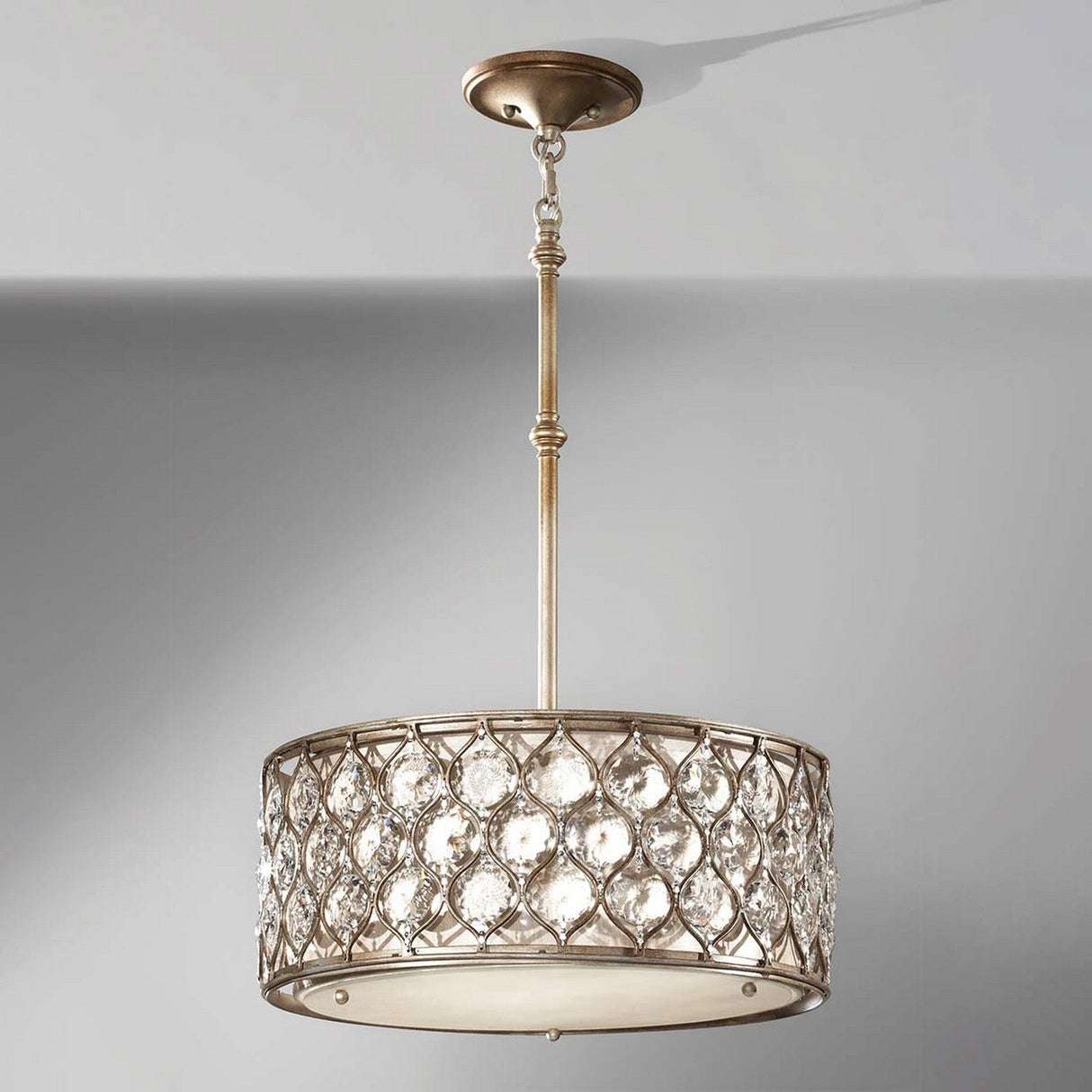 The Lucia 3 Light Chandelier - Burnished Silver is a decorative ceiling fixture with a drum-shaped shade, featuring a metallic frame in burnished silver and adorned with large, clear Bauhinia crystal accents. It elegantly hangs from a single rod against a plain, light gray backdrop.