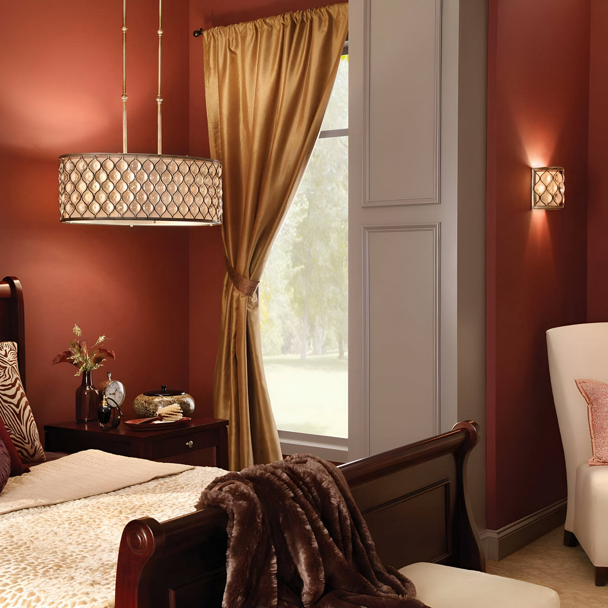 A cozy bedroom featuring dark wood furniture, a bed adorned with patterned pillows, and a plush brown blanket. Warm red walls complement a window draped with gold curtains. A Lucia 3 Light Large Chandelier in Burnished Silver hangs stylishly above the bedside table, adding an elegant touch to the decor.