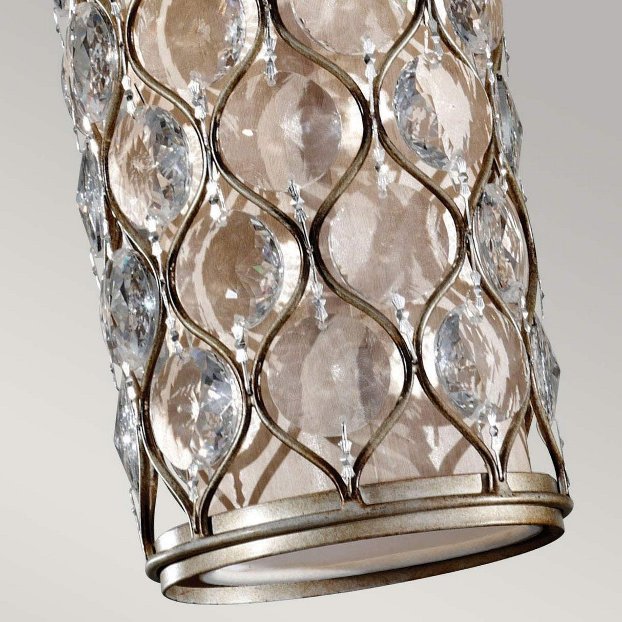 A close-up of the Lucia 1 Light Mini Pendant in Burnished Silver features a cylindrical silhouette with an intricate metal lattice design adorned with Bauhinia Crystals. The large, clear accents lend a luxurious and sparkling appearance against a neutral background.