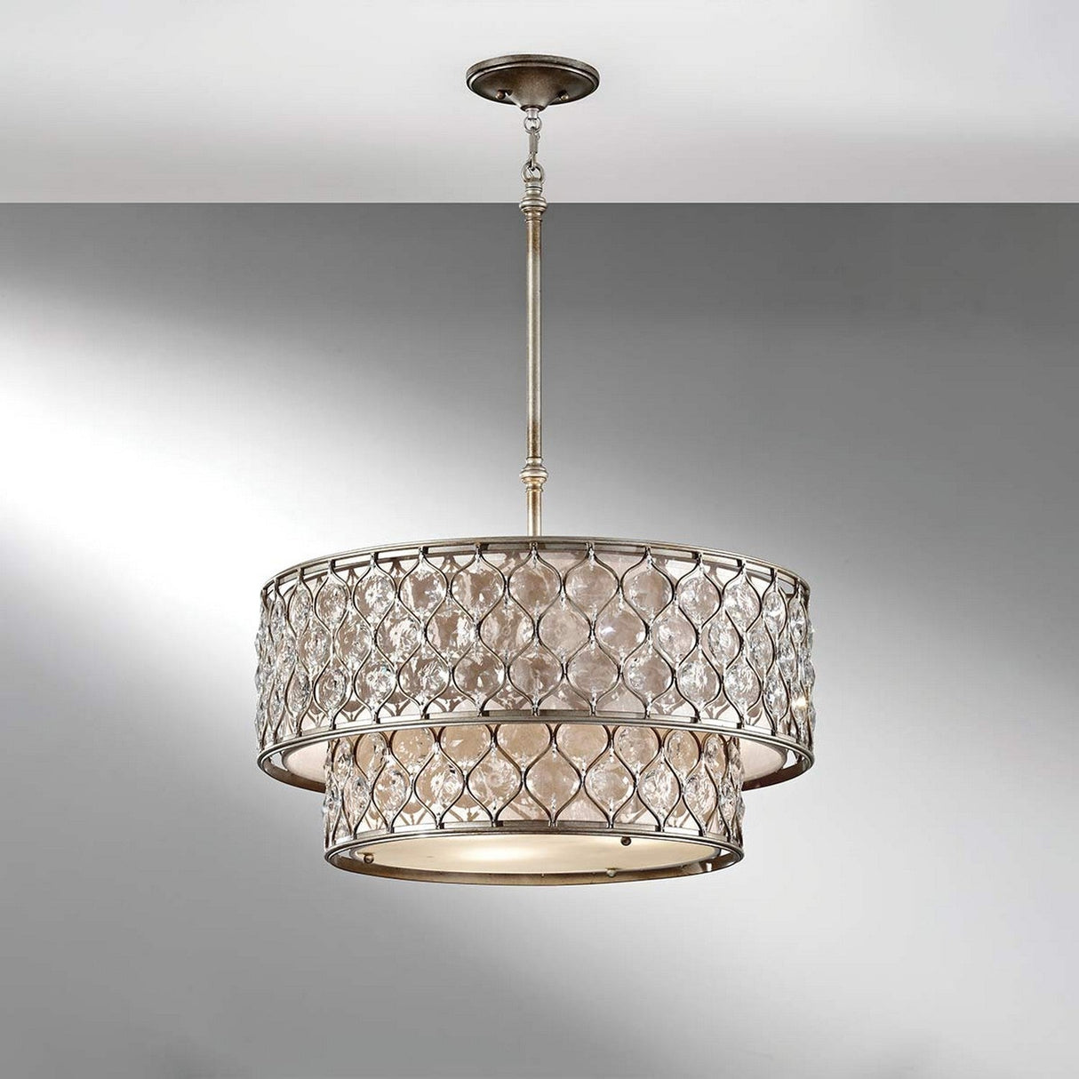 The Lucia 6 Light Chandelier in burnished silver is suspended from the ceiling, showcasing a contemporary pendant style with a cylindrical design. It boasts a lattice-patterned outer frame and frosted glass inner layer, providing a soft, diffused glow against the white and gray wall backdrop.