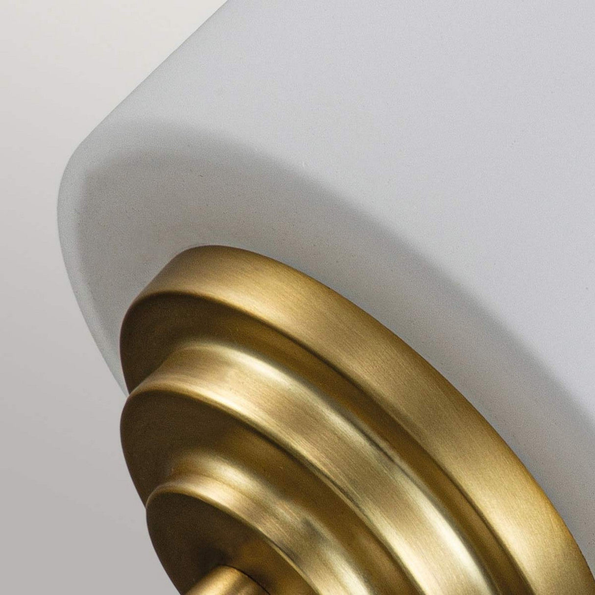 Close-up of the Malibu 1 Light Wall Light in brushed brass, showcasing its elegant combination of a matte white, opal-etched glass shade and tiered base. The simple yet sophisticated design effortlessly merges contemporary and classic elements against a neutral backdrop.