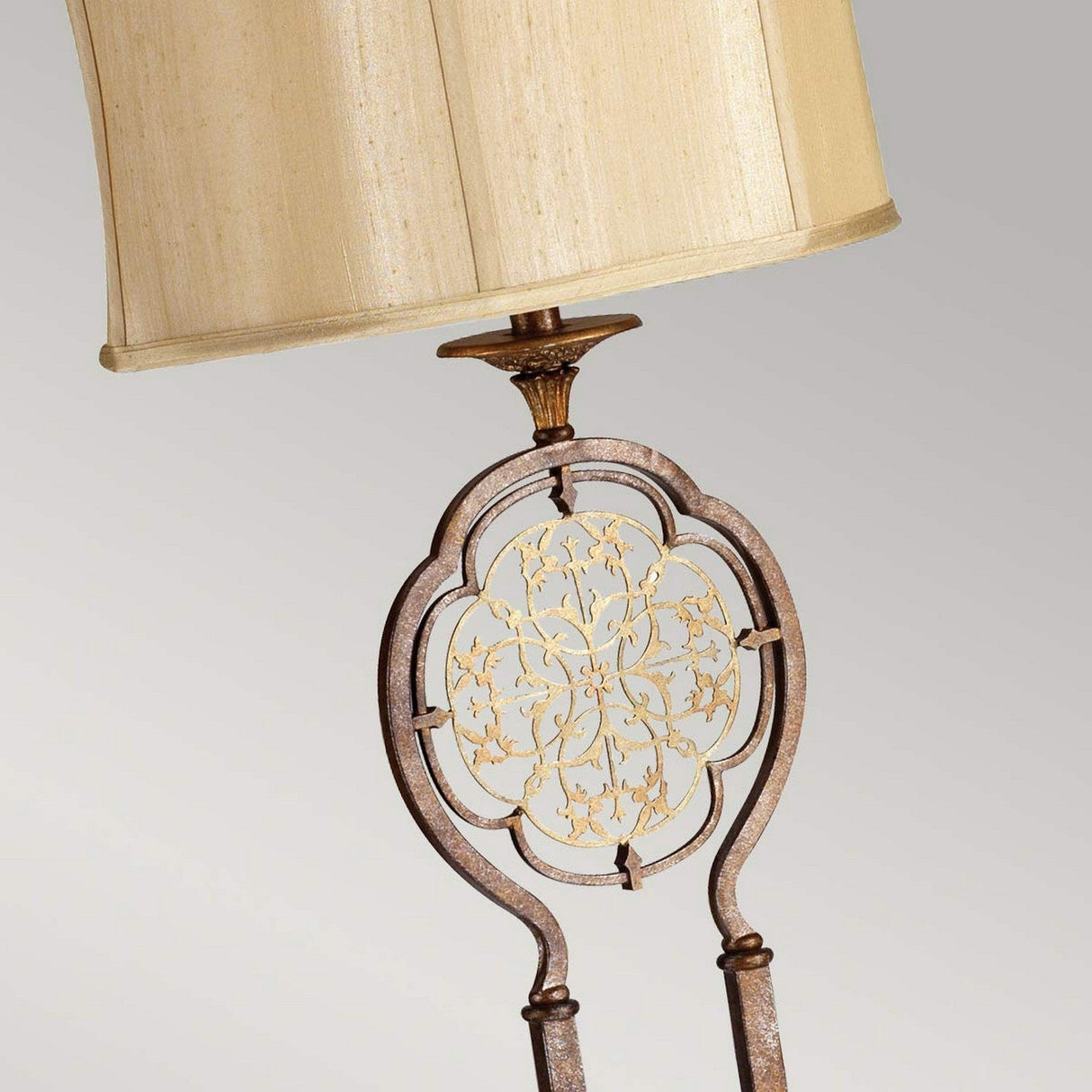 The Marcella 1 Light Floor Lamp - Bronze showcases an antique bronze finish with a gold floral design and filigree details on the stem, complemented by a beige, curved lampshade. This lamp features intricate metalwork and a decorative base set against a neutral background, exuding vintage glamour.