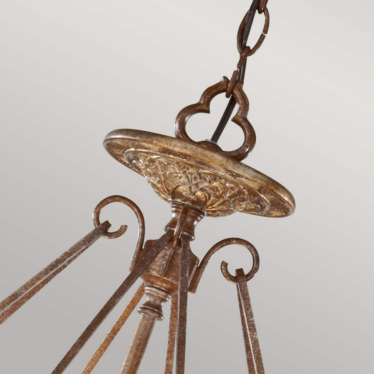 Close-up of the Marcella 3 Light Chandelier in Bronze, showcasing an ornate antique-style design with a decorative metal canopy and curved arms. The fixture, offering ambient illumination with its rustic patina finish, hangs elegantly from a sturdy chain against a gray background.