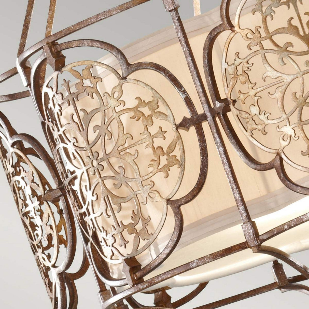 Close-up of the Marcella 3 Light Chandelier in a bronze ornate filigree design featuring intricate floral patterns. This design surrounds a cylindrical cream-colored lampshade, creating an elegant and decorative appearance with ambient illumination.