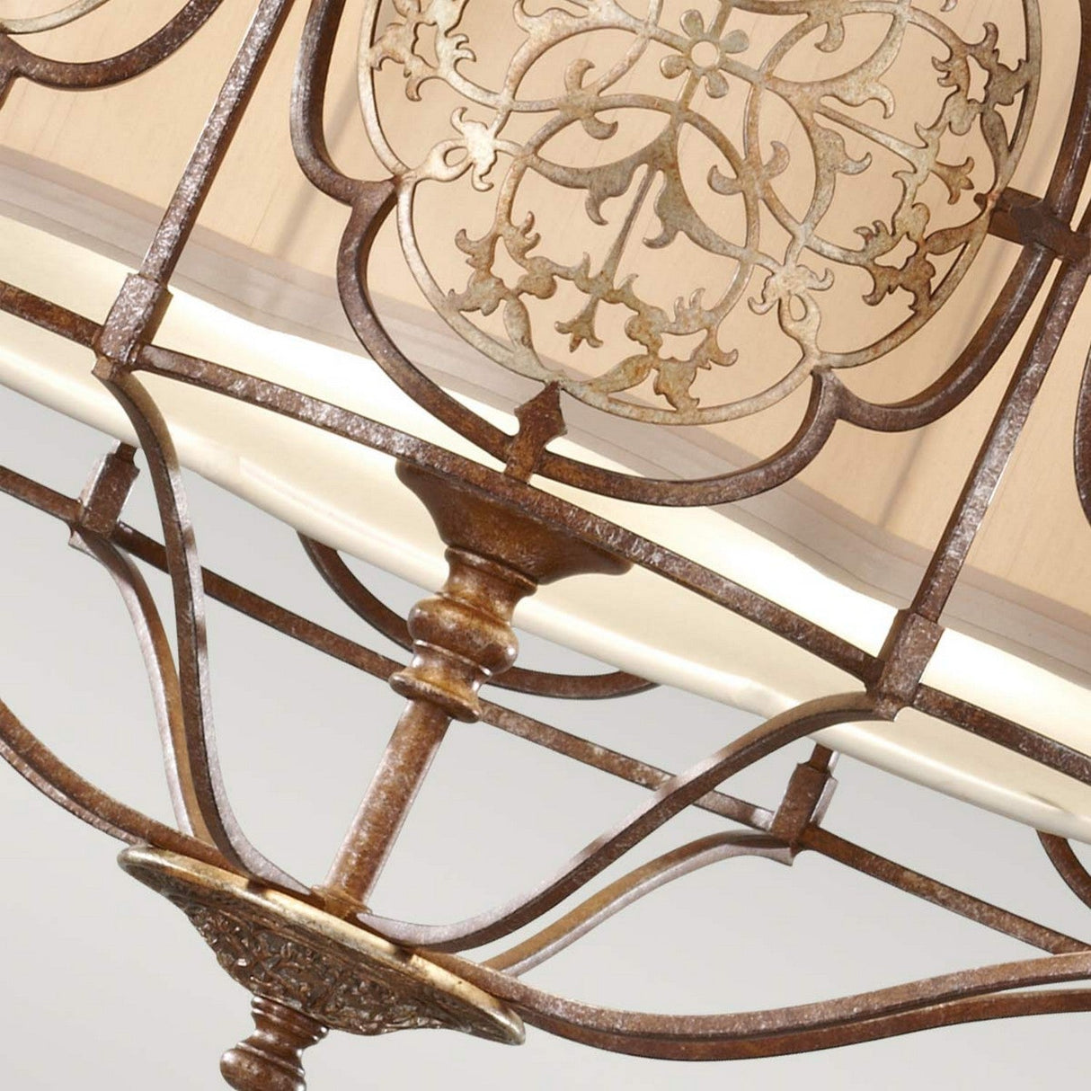A close-up of the Marcella 3 Light Chandelier - Bronze highlights its intricate scrollwork and floral patterns. The bronze filigree design features a rustic finish, beautifully complemented by the ambient glow of a cream-colored shade in the background.