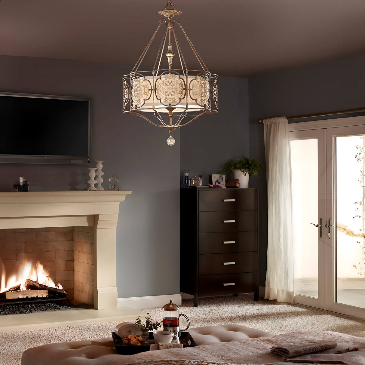 A cozy living room features ambient illumination, showcasing the Marcella 3 Light Chandelier in Bronze and a dark wood dresser. Glass doors lead outside, while a French press with coffee adorns the cushioned ottoman in the foreground.