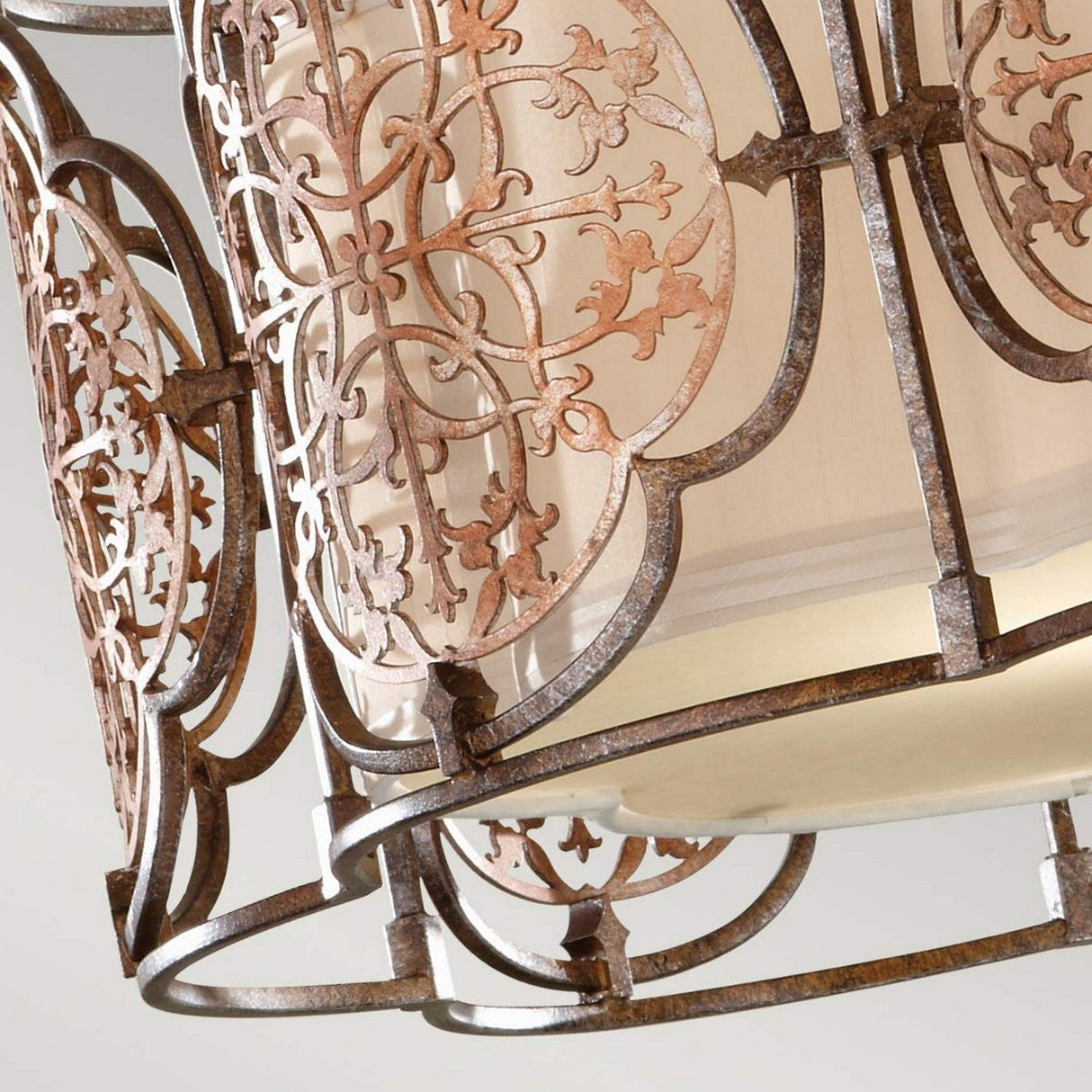 A close-up of the Marcella 3 Light Semi Flush in bronze reveals an elegantly exotic metal lantern with an ornate floral pattern and a weathered finish. Its captivating design allows glimpses of the beige fabric shade.