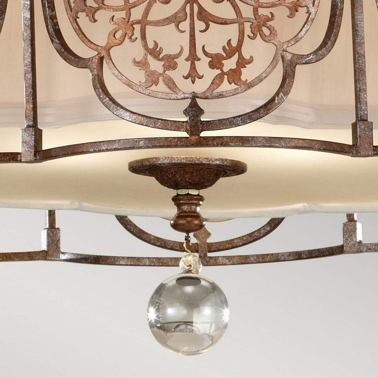 A close-up of the Marcella 3 Light Semi Flush chandelier showcases intricate wrought iron scrollwork and a central crystal sphere. This chandelier radiates exotic elegance with its light, elegant design, finished in bronze and complemented by cream-colored shades.