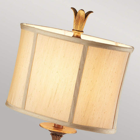 The Marcella 1 Light Table Lamp in bronze features a beige faux silk lampshade with vertical panels and intricate filigree details, crowned with a gold feather-shaped finial. This sophisticated lamp stands elegantly against a light gray background.