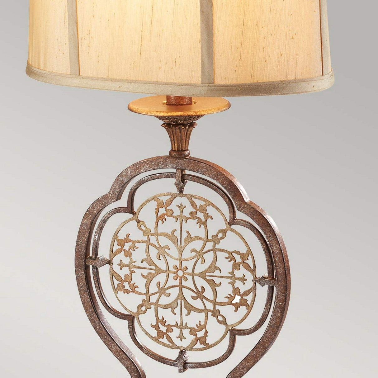 The Marcella 1 Light Table Lamp - Bronze is a decorative piece featuring an ornate metal base with intricate filigree details and elegant floral patterns. It is complemented by a light brown cylindrical faux silk shade, all set against a neutral gray background.