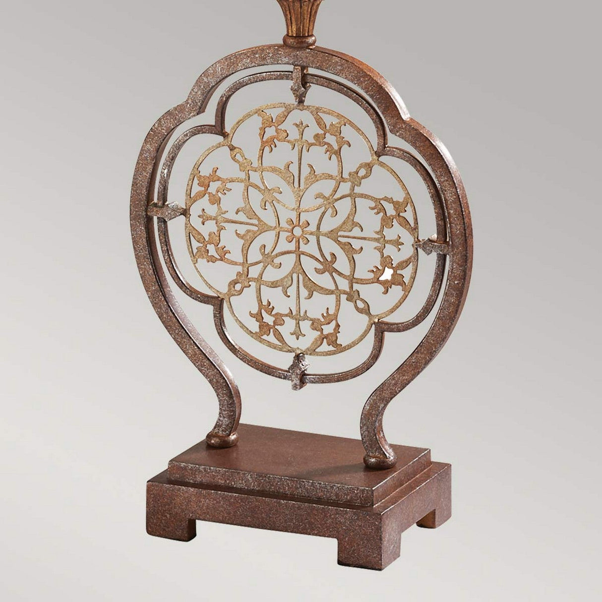 The Marcella 1 Light Table Lamp - Bronze features an ornate metal sculpture with filigree details and a floral and vine design at its center, set on a dark rectangular base. Its intricate pattern is surrounded by a symmetrical, curved border against a solid gray background.