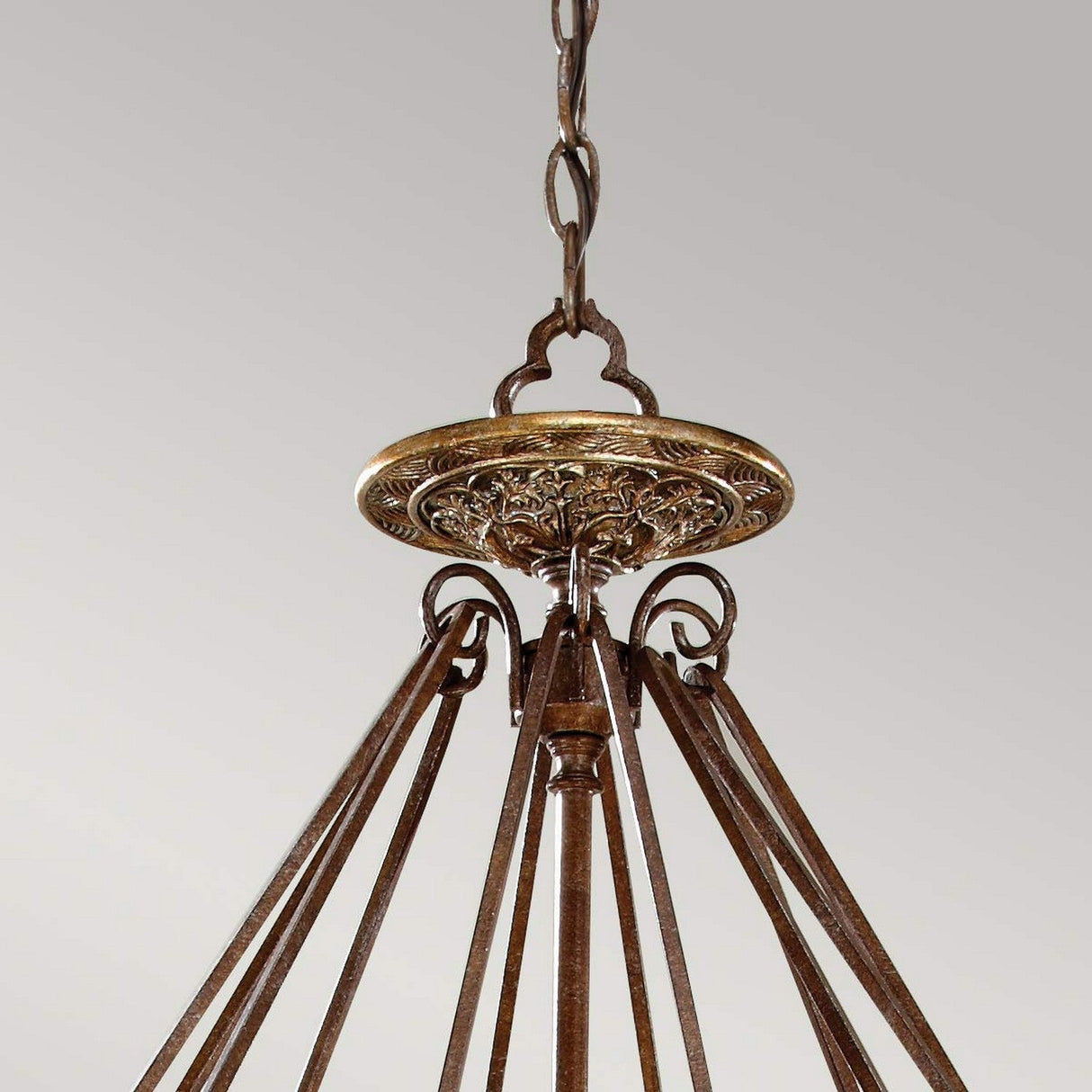 The Marcella 4 Light Chandelier - Bronze showcases luxury lighting with a decorative circular base and elegantly curved metal arms, all gracefully suspended by a chain against a gray background.