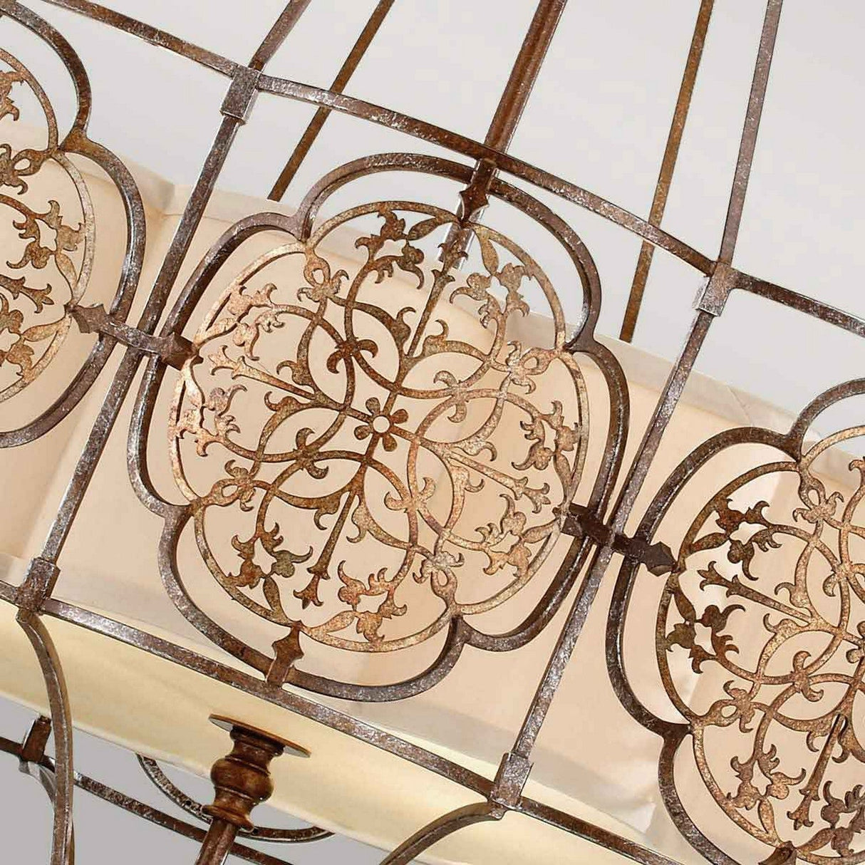 Close-up of the Marcella 4 Light Chandelier - Bronze, showcasing its ornate metal design with intricate floral patterns on a circular frame. The rustic bronze finish and the luxurious lighting feature a beige fabric shade that is partially visible in the background, enhancing its antique charm.